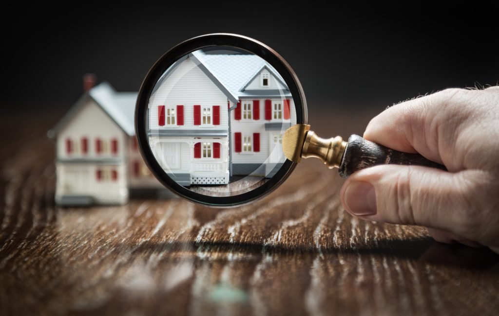 Hand Holding Magnifying Glass Up To Model Home on Reflective Wooden Surface.